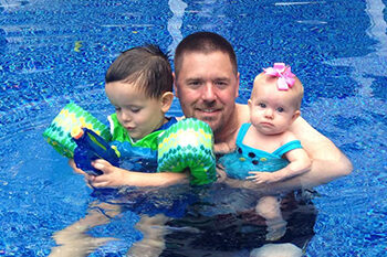 A man holds an infant and a toddler in his arms in a swimming pool.
