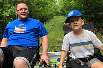 A man and a child ride downhill through a wooded location on a cart along rails.