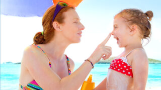 Red haired woman putting sunscreen on her daughter at the beach to call attention to expert care for skin cancer.