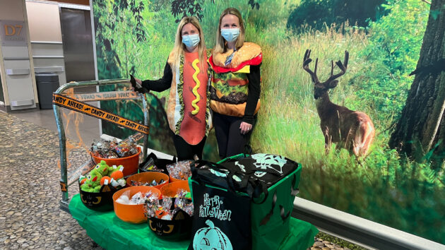 Ronald McDonald charity volunteers (dressed as a hot dog and cheeseburger), stand next to a cart full of Halloween candy, trick or treat bags, and toys