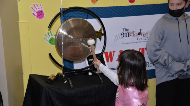 Young girl, wearing a superhero cape, rings a gong in the Melodies Center to celebrate finishing treatment.