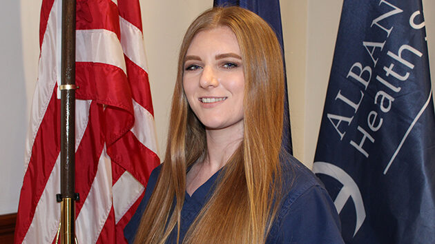 Megan Ruddock-Rees, RN, MSN, posing in front of any American flag and the Albany Med Health Systems flag