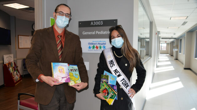 Dr. Stephen de Waal Malefyt (left) and Mrs. Queen of the World, New York, Enza Castle (right) holding children's books outside the General Pediatrics Office
