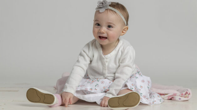 A baby wearing a white dress sits and smiles at someone out of frame