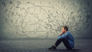 A man in a blue shirt and black pants sits on the ground, facing left, in front of a background of lines, symbols, and words, that attempt to characterize his thoughts related to stress and mental health.