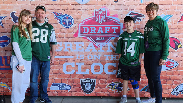 Kyle and his family, wearing NY Jets shirts, stand in front of a sign that says NFL Draft