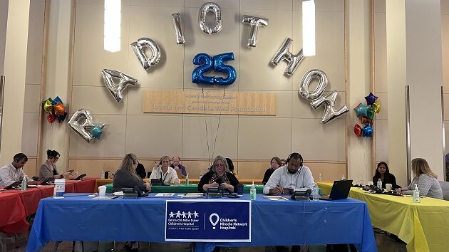 People sitting at tables with multi-colored tablecloths, answering phones. Balloons hang over head that say "Radiothon 25"