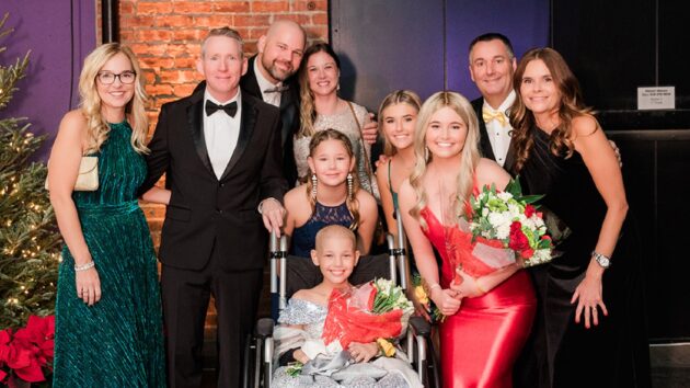 A group of people, dressed in formal attire, posing for a picture. Two young girls, one in a wheelchair, are holding flowers. They are the guests of honor at the gala.