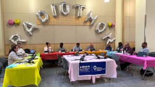 Radiothon Volunteers sitting at tables in front of phones. "Radiothon" balloons hang overhead.