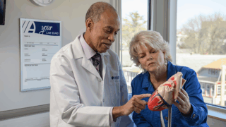 A doctor points to a portion of a heart model while discussing results with a seated patient.
