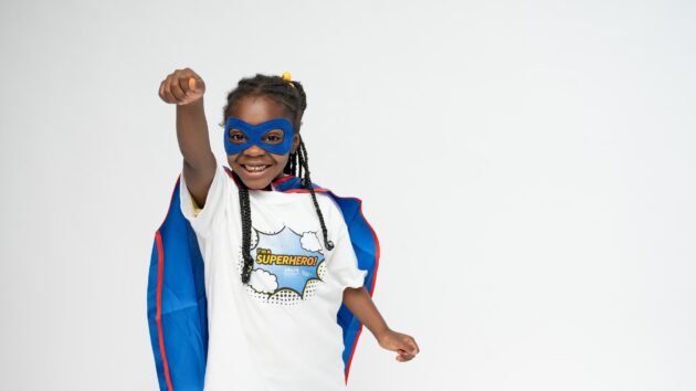 A young girl poses like a superhero in a cape and eye mask