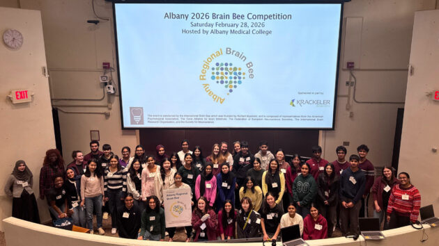 A large group of students standing at the front of a lecture hall in front of a screen with information about the 2026 Albany Regional Brain Bee
