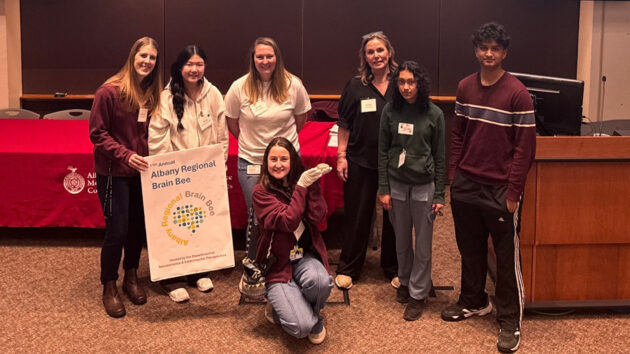 The top three scorers at the 2026 Albany Regional Brain Bee standing at the front of a lecture hall with Albany Brain Bee committee members.