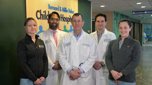 Three doctors in white coats, a Physician Assistant, and a Nurse Practitioner pose for a group picture in the children's hospital hallway