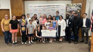 A group of adults and kids gather for a check presentation. Little kids in the front row are holding the big check.