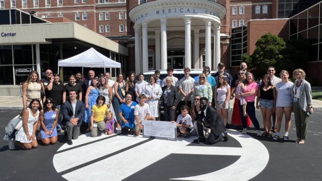 A large group of adults and children gathered in front of the Albany Medical Center Pillars for a check presentation with Hyundai Hope on Wheels. Young kids in the front are holding the big check.