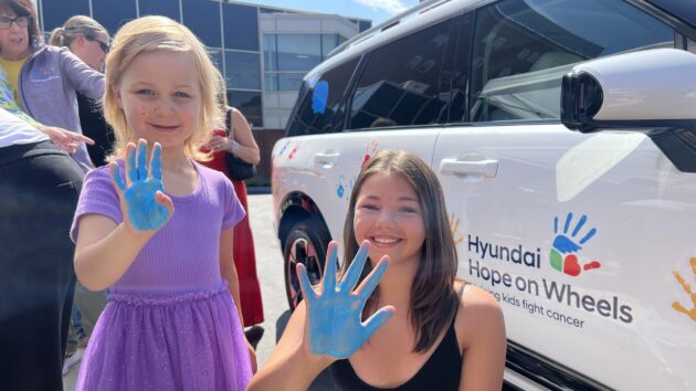 Two girls are holding up their hands that are painted blue. They're standing in front of a white car covered in painted handprints, as part of the Hyundai Hope on Wheels event.