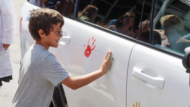 A young boy is pressing his painted handprint on a white car that's covered in painted handprints, as part of the Hyundai Hope on Wheels event.