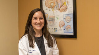 Neurosurgeon Dr. Brannan O'Neill wearing a white coat and standing in front of a poster about the brain