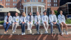 Group of eight students from the 2026 Clinical Immersion Program sit on a ledge wearing white coats outside Albany Medical Center's Pillars entrance