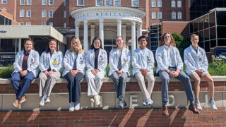 Group of eight students from the 2026 Clinical Immersion Program sit on a ledge wearing white coats outside Albany Medical Center's Pillars entrance